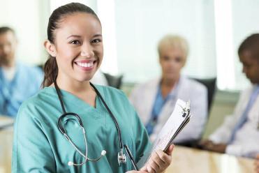 A portrait of a woman in green scrubs carrying a medical chart with other medical professionals in the distant background. 