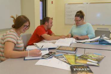 Three students sit at a table filled with math books and calculators, with the center student pointing out something in an open textbook before them.